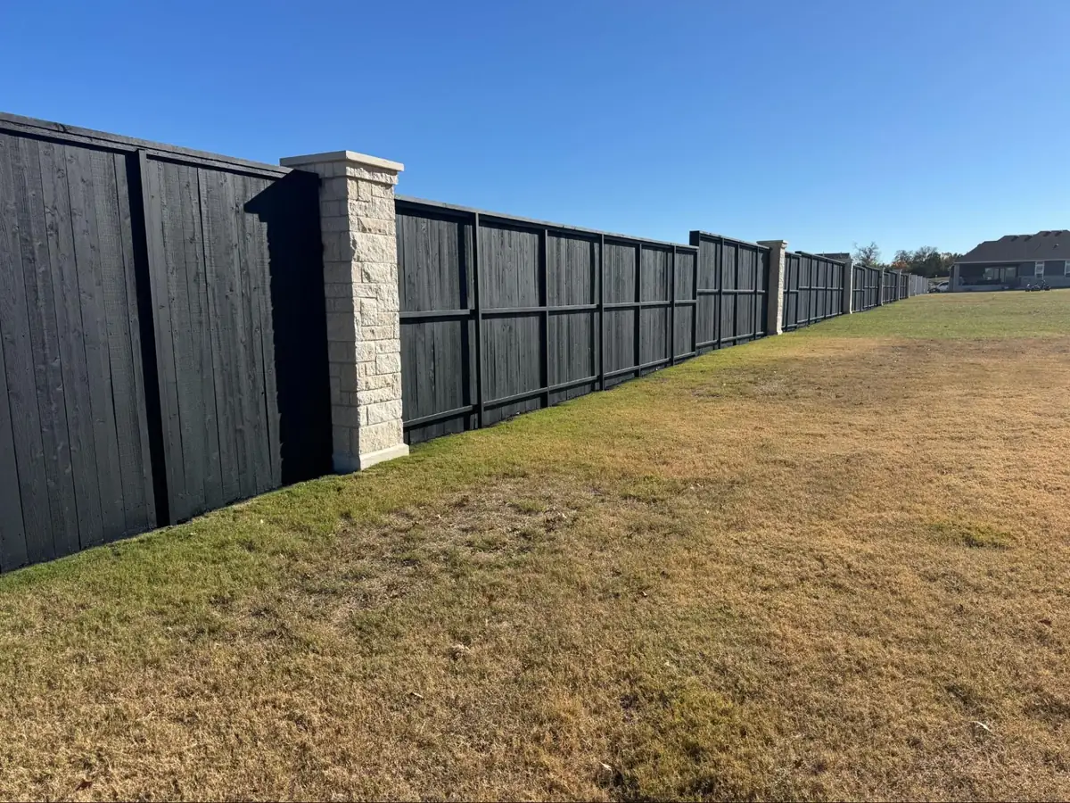 A long, modern black-stained wood privacy fence seamlessly integrated with light-colored stone masonry columns along an open property line under a clear blue sky.