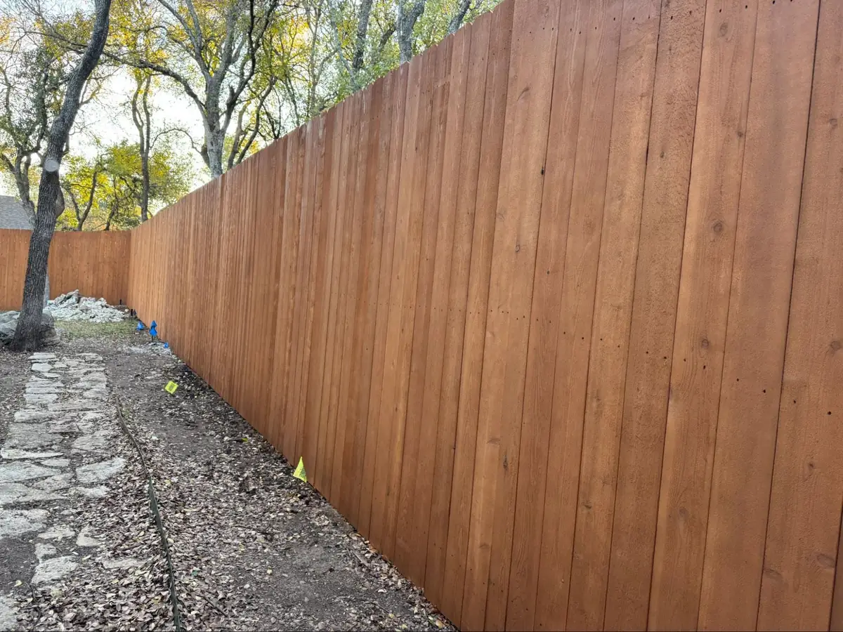 A tall wooden privacy fence runs alongside a dirt path with scattered leaves, bordered by trees with yellowing foliage.