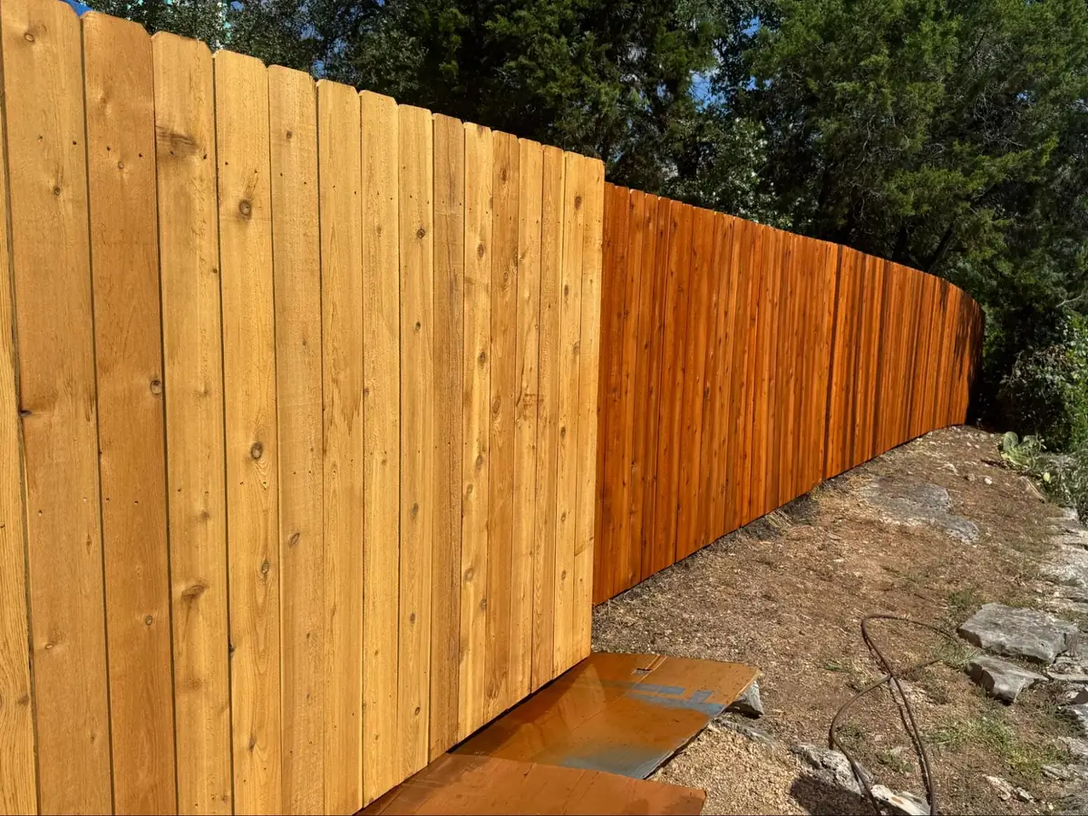 A wooden fence with the left side unstained and the right side treated with a darker wood stain, surrounded by trees and dirt ground.